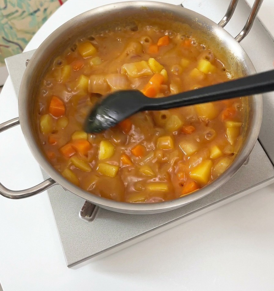 Czarna łopatka mieszająca gęsty, bulgoczący sos curry z widocznymi kawałkami warzyw w srebrnym garnku.