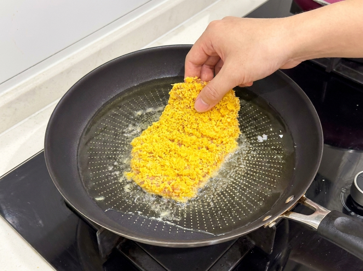 Panierowany kotlet z kurczaka wkładany na patelnię z gorącym, lśniącym olejem.