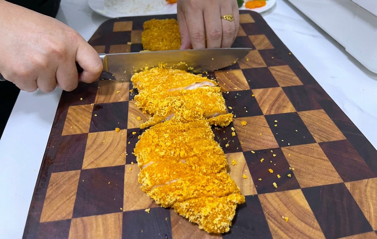 Osoba krojąca dużym nożem chrupiący, panierowany kotlet z kurczaka na ciemnej, drewnianej desce.
