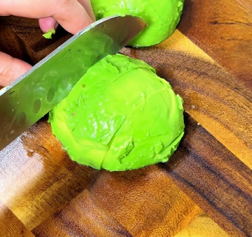 Zbliżenie na dłonie krojące dojrzałe awokado w równą kostkę na drewnianej desce kuchennej.