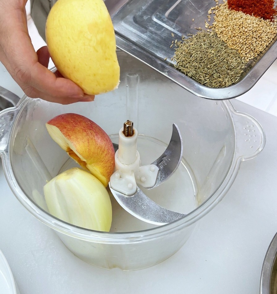 Kawałki świeżych owoców wkładane do misy robota kuchennego wraz z tacą gotowych przypraw.