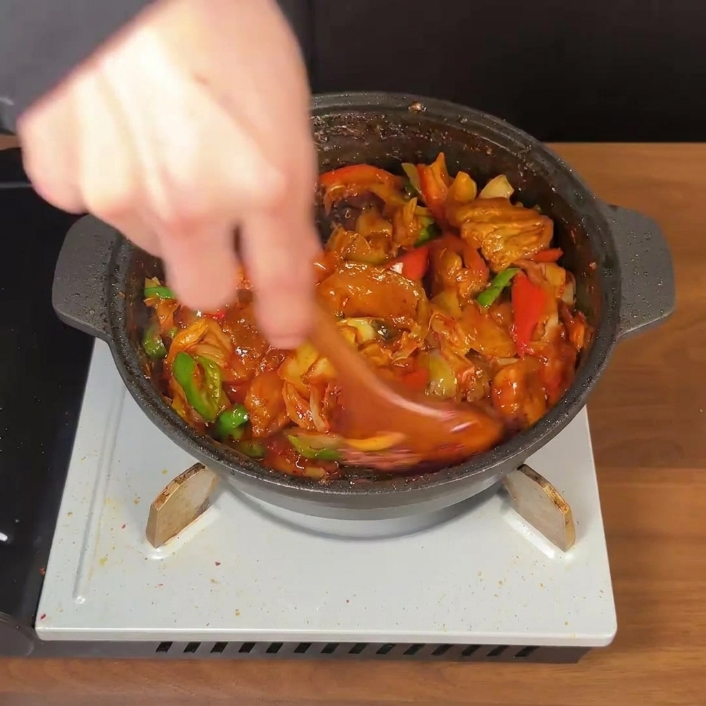 Drewniana łyżka energicznie mieszająca boczek, kimchi i papryczki w gęstym czerwonym sosie.