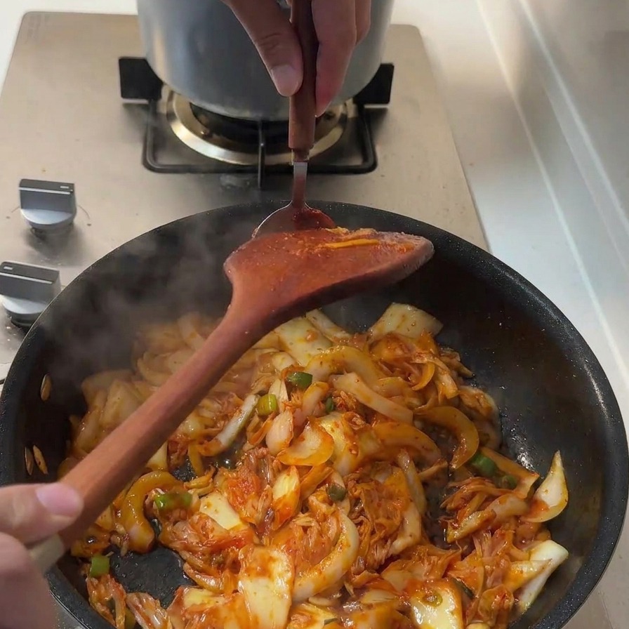 Łyżka gęstej, czerwonej pasty gochujang dodawana do składników na patelni.