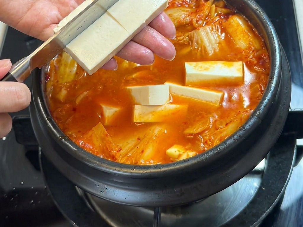 Osoba krojąca kostkę białego tofu bezpośrednio nad parującym garnkiem ostrego gulaszu.