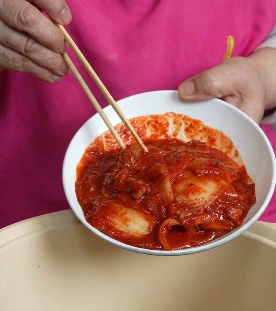 Biała miska pełna czerwonego, soczystego kimchi przechylana nad garnkiem z boczkiem.