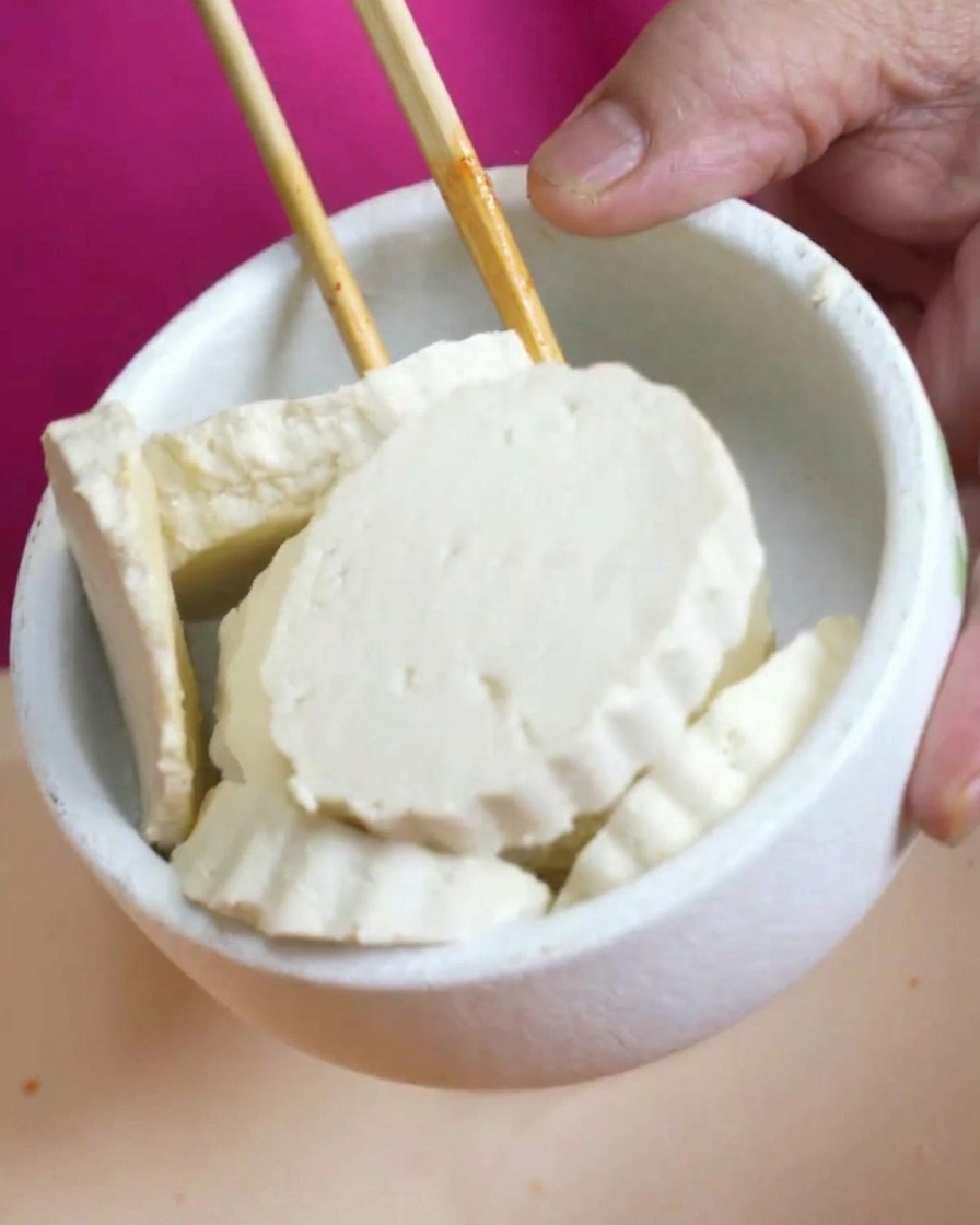 Mała biała miseczka z grubymi plastrami białego tofu trzymana nad parującym garnkiem.