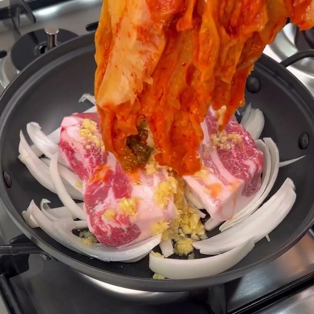 Ręka kładąca dużą porcję czerwonego, sfermentowanego kimchi na surowym boczku.