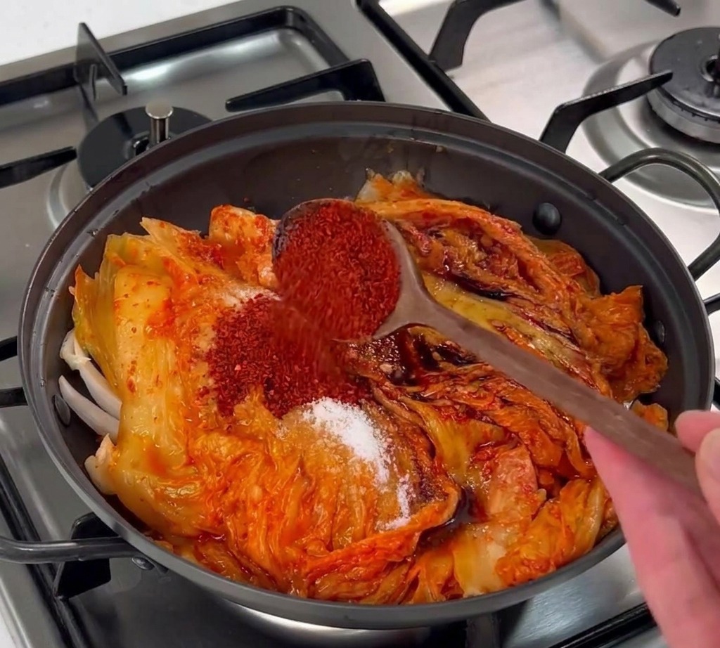 Drewniana łyżka wsypująca czerwone koreańskie płatki chili na warstwę kimchi w garnku.