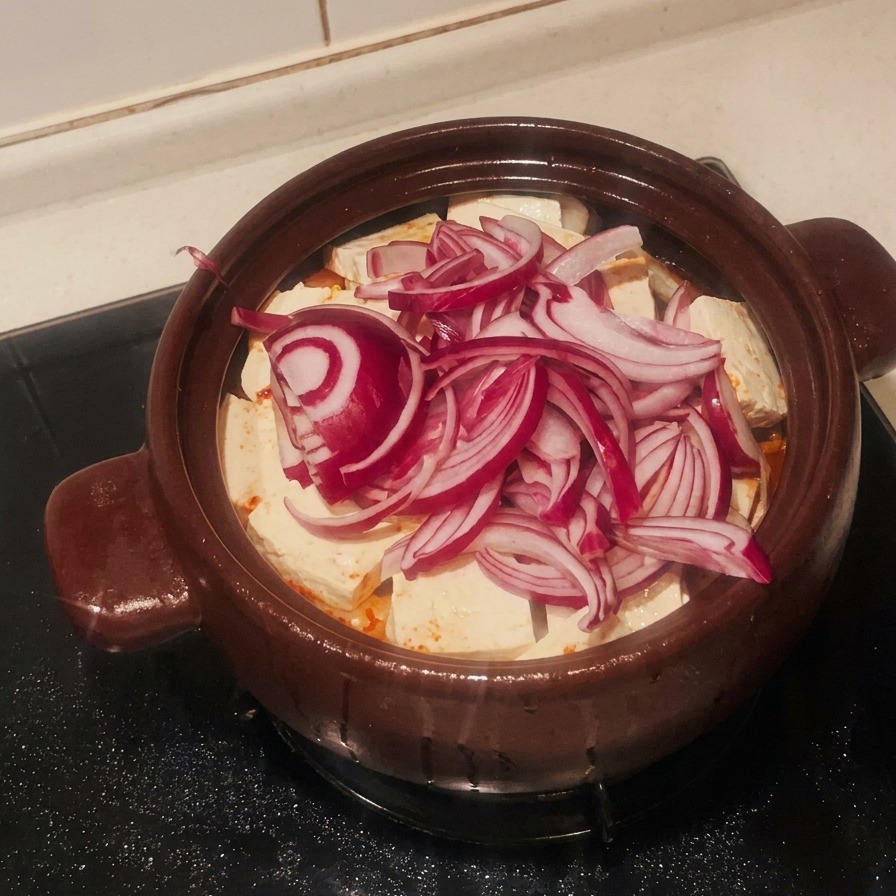 Warstwa pokrojonej czerwonej cebuli przykrywająca tofu w naczyniu glinianym.
