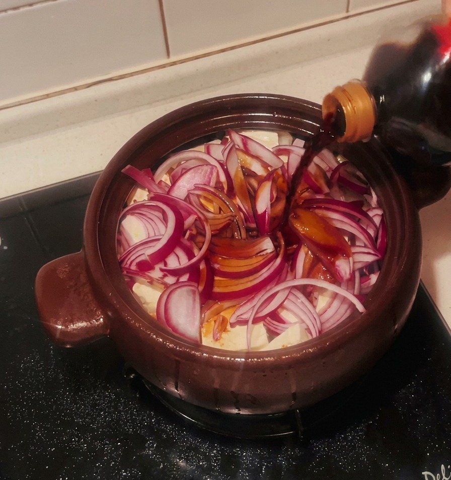 Ciemny sos sojowy wlewany z butelki na warstwę cebuli i tofu w brązowym naczyniu glinianym.
