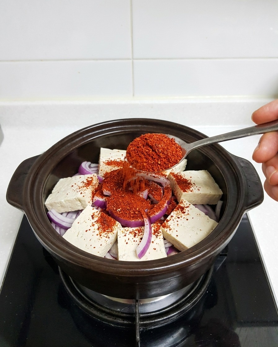Metalowa łyżka nakładająca jaskrawoczerwony proszek chili na tofu i cebulę w naczyniu.