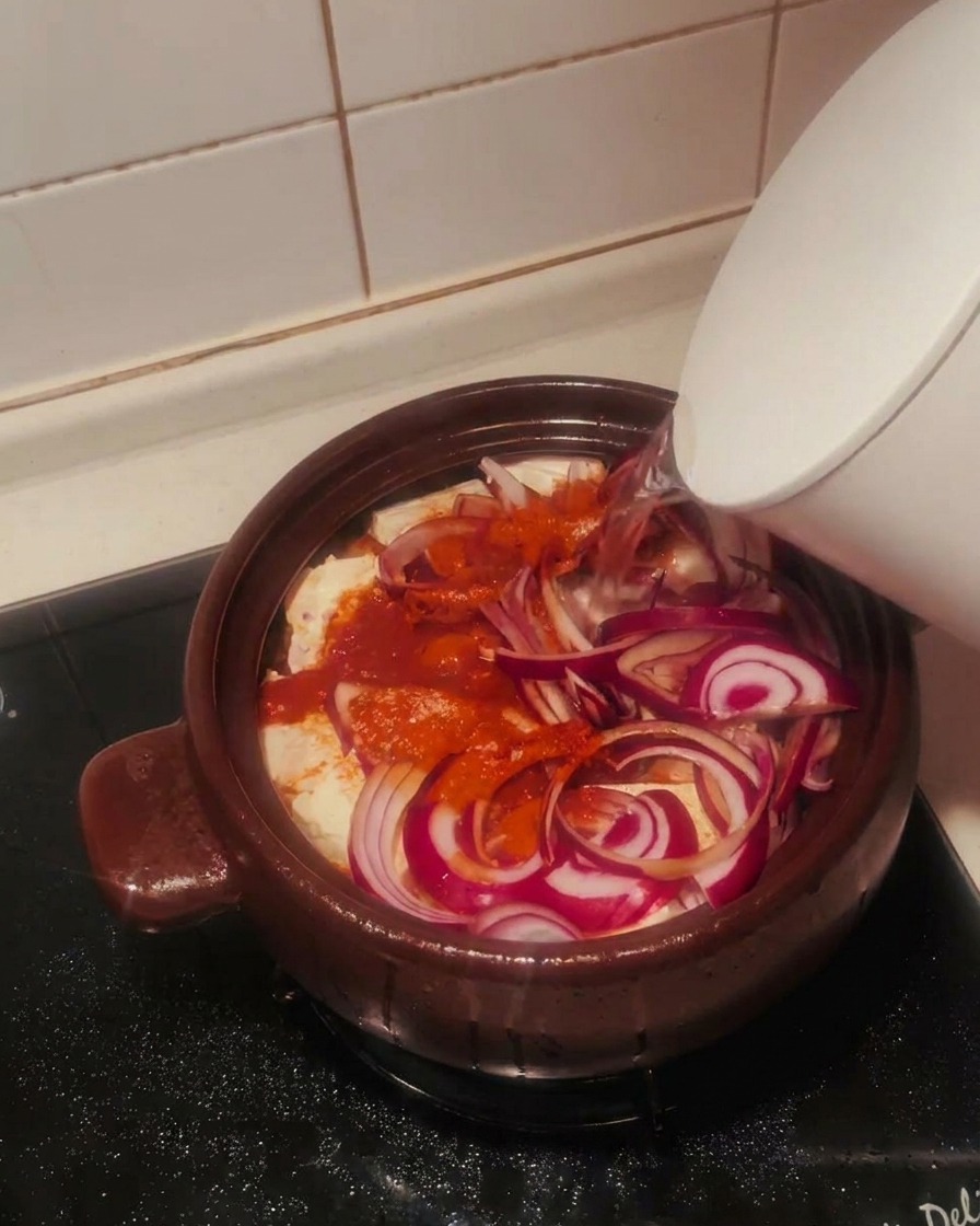 Woda wlewana z białego czajnika do naczynia wypełnionego kimchi, tofu, cebulą i przyprawami.