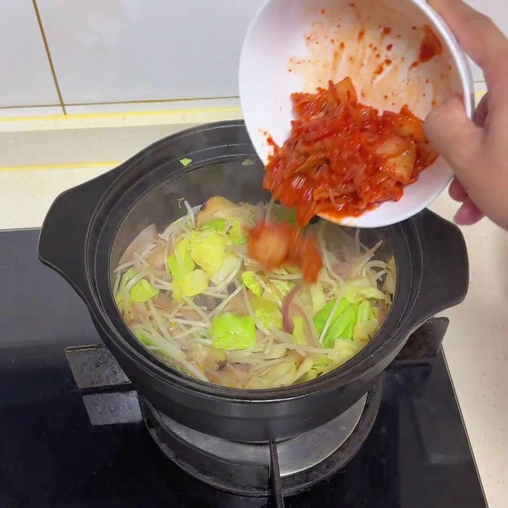 Jasnoczerwone posiekane kimchi wsypywane z białej miski do garnka z podsmażonymi warzywami.