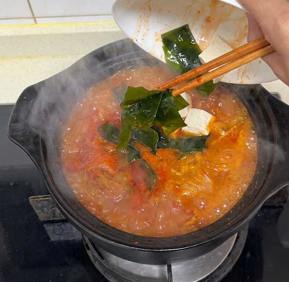 Dłoń wrzucająca kawałki ciemnozielonych wodorostów kombu do parującego garnka z bulionem kimchi.
