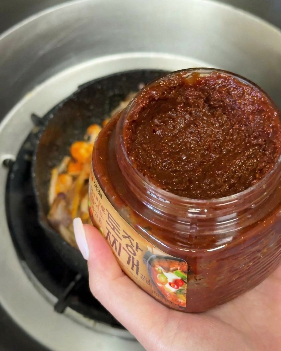 Dłoń trzymająca łyżkę gęstej, ciemnobrązowej pasty Doenjang nad patelnią z podsmażonymi warzywami.