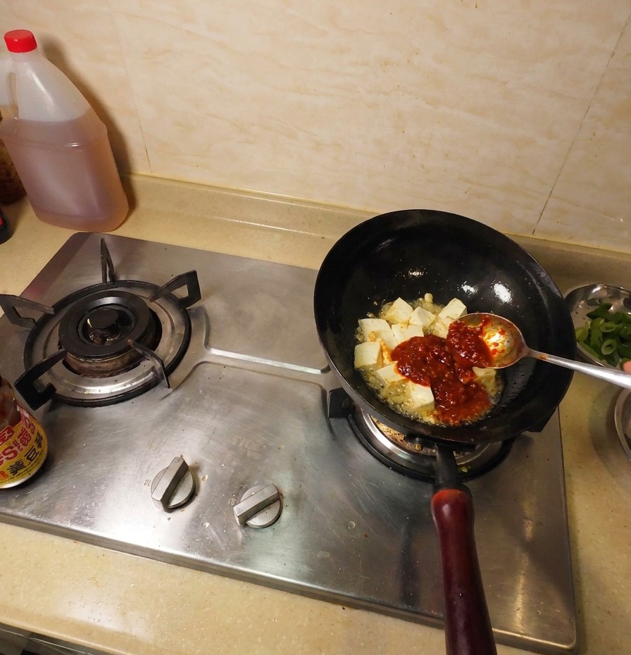 Ciemnobrązowa pasta sojowa na metalowej łyżce dodawana do woka obok tofu i sosu chili.