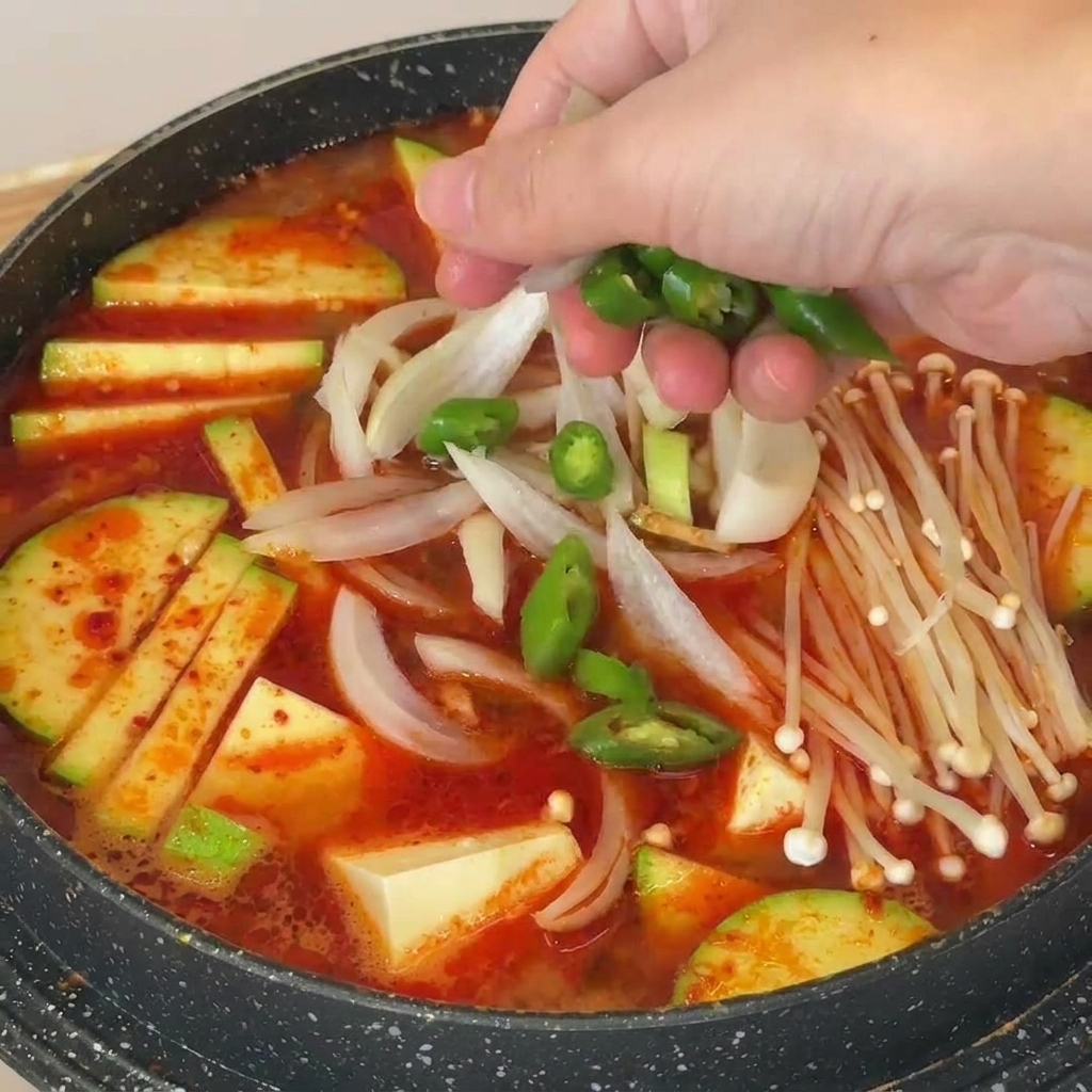 Ręka posypująca plastrami zielonego chili koreańską zupę z pasty sojowej z grzybami, tofu i cukinią.