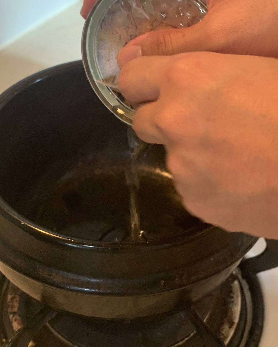 Wlewanie aromatycznego oleju z puszki tuńczyka do czarnego ceramicznego garnka na kuchence.