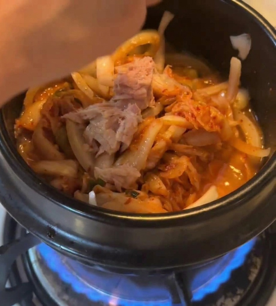 Kawałki tuńczyka z puszki dodawane do garnka z kimchi na kuchence gazowej.