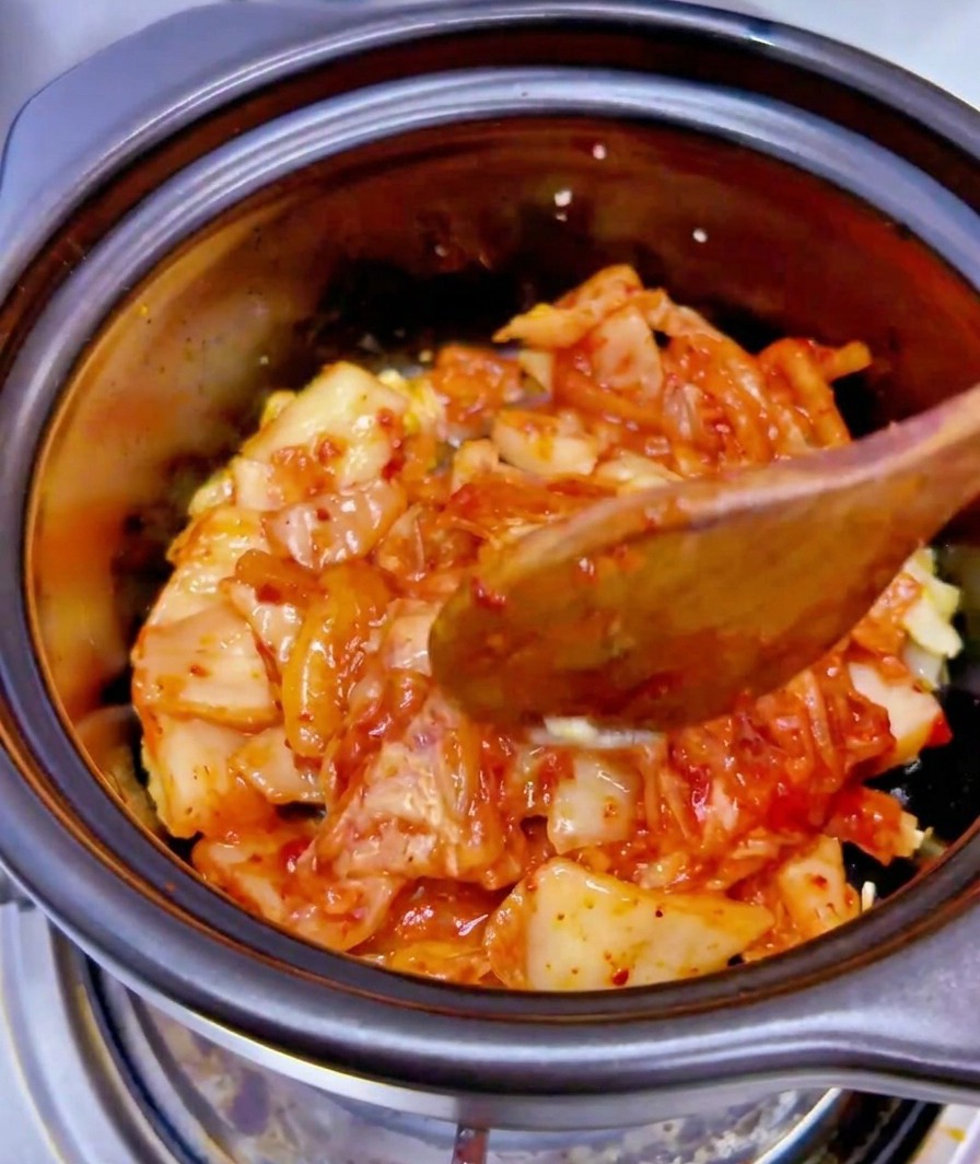Płyn z puszki tuńczyka wlewany bezpośrednio na mieszankę kimchi i czosnku w czarnym garnku.