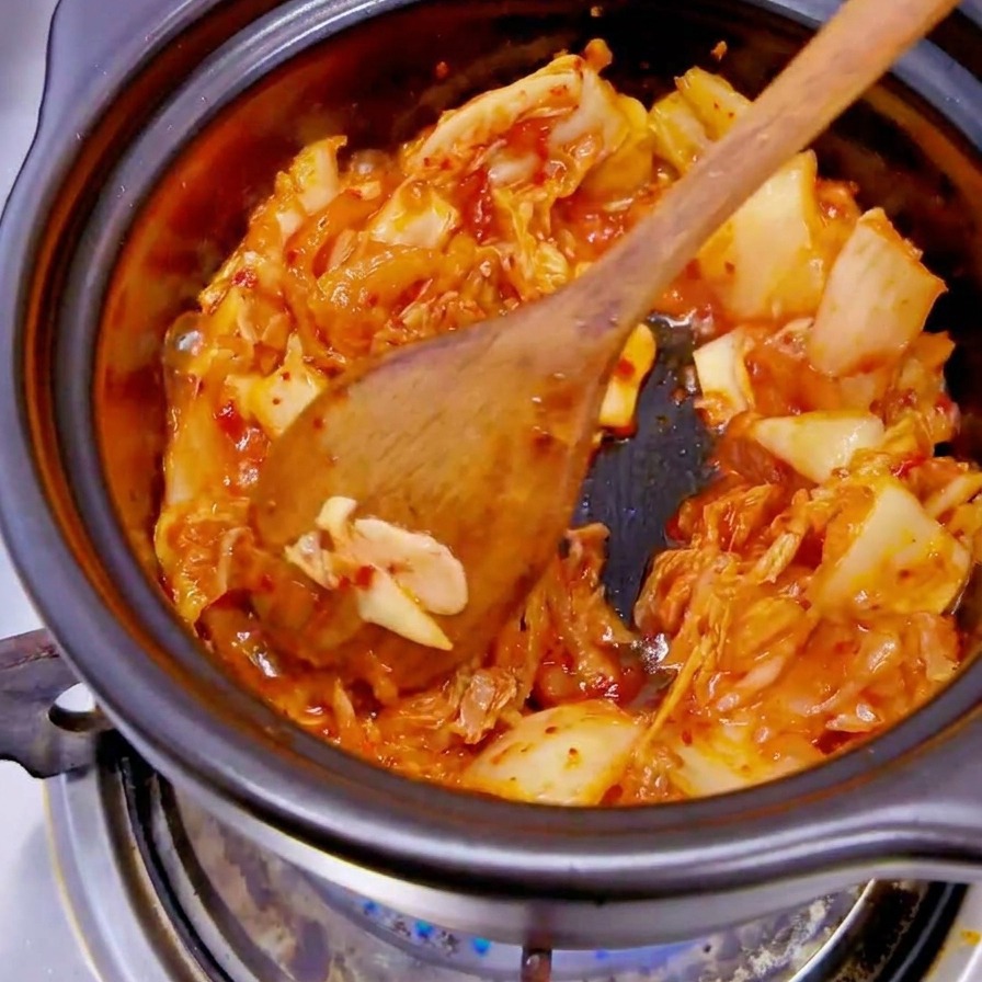 Ręka w czarnej rękawiczce wrzucająca plasterki białej cebuli do garnka z czerwonym kimchi.