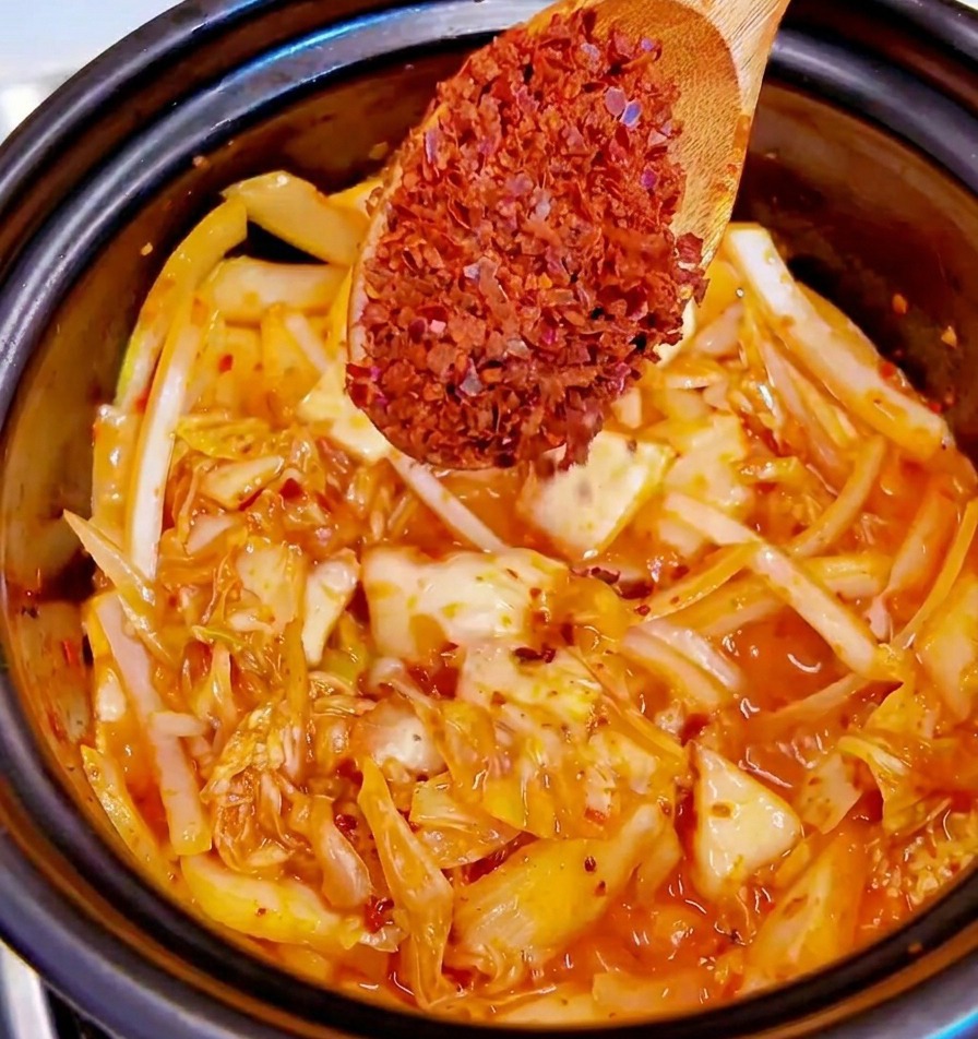 Drewniana łyżka z gruboziarnistymi płatkami gochugaru nad garnkiem z podsmażanym kimchi i cebulą.