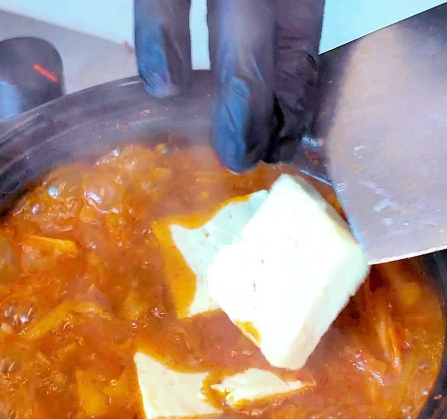 Plastry białego tofu wkładane delikatnie do wrzącej, czerwonej zupy kimchi za pomocą łopatki.