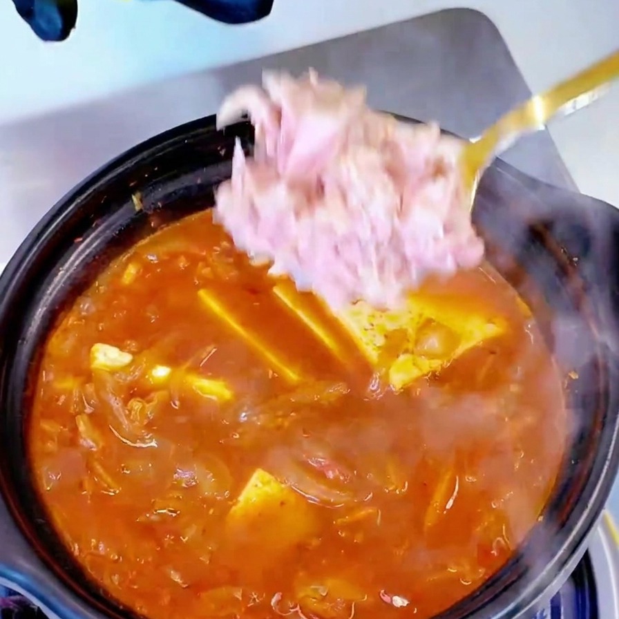 Kawałki tuńczyka z puszki nakładane łyżką do garnka z wrzącą zupą kimchi i tofu.