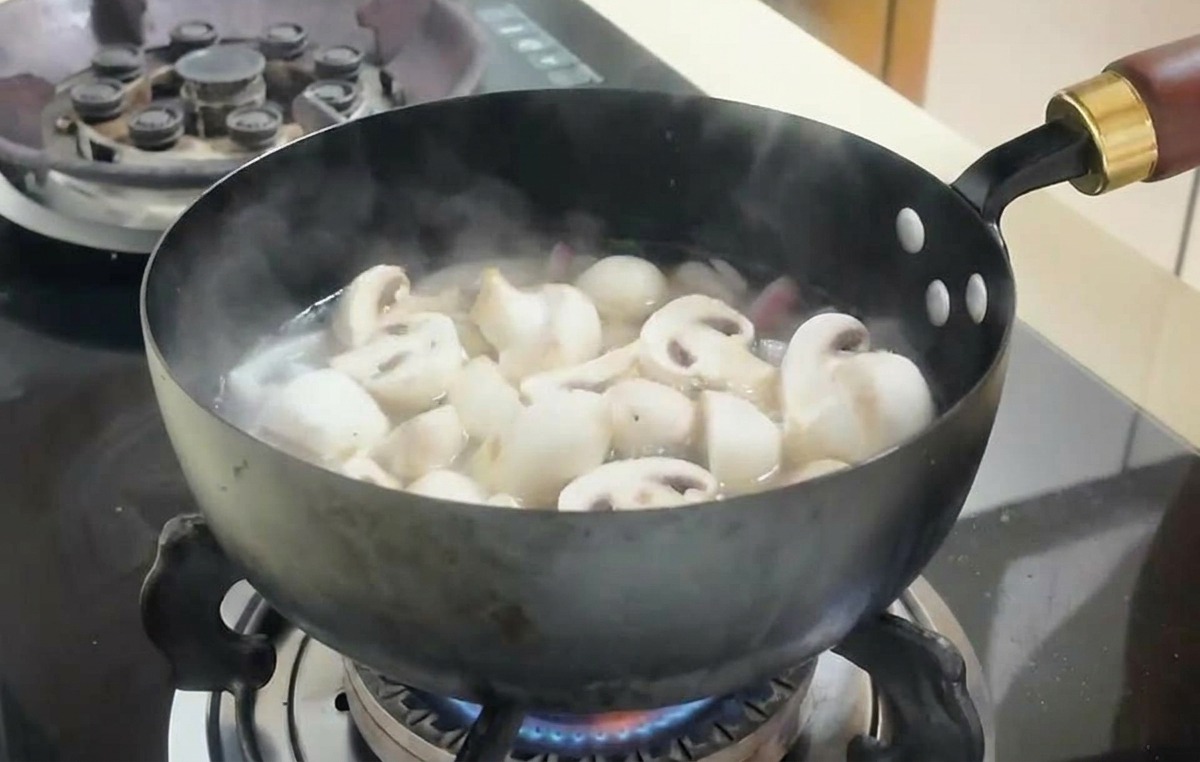 Plasterki białych pieczarek i kostki tofu gotujące się w bulionie na ciemnej patelni.