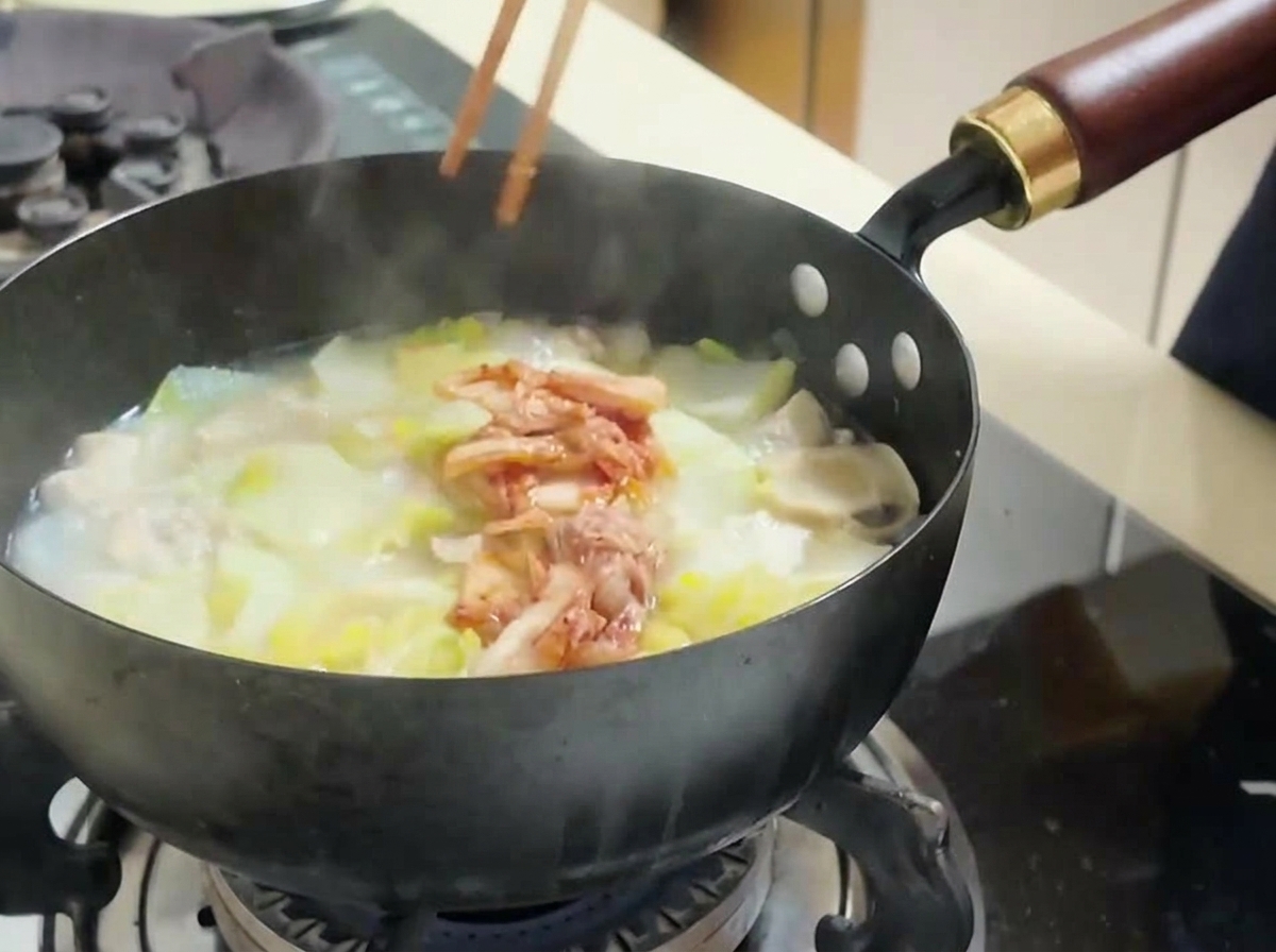 Osoba dodająca pałeczkami porcję czerwonego kimchi do garnka z zupą, tofu i kapustą.