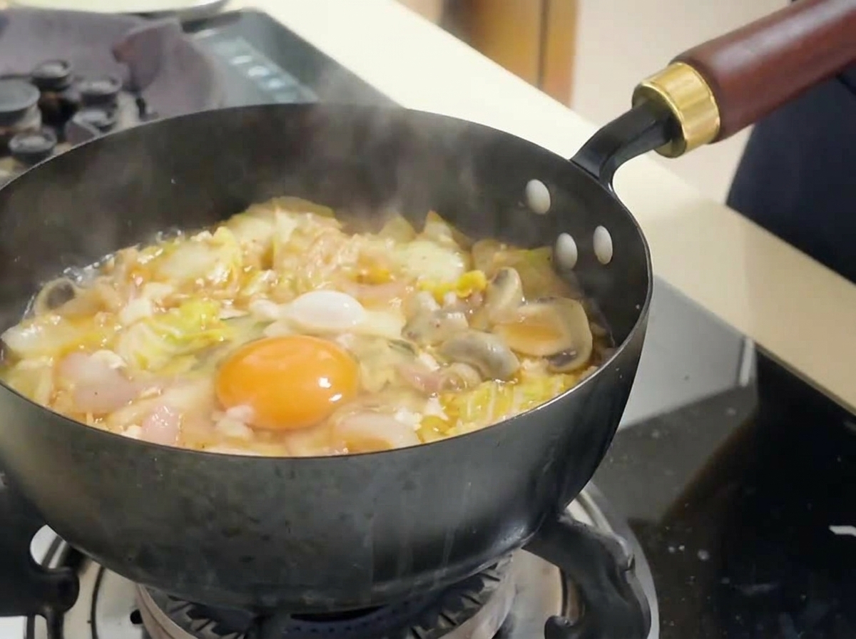 Surowe żółtko jajka na środku gotującej się czerwonej zupy kimchi, otoczone kapustą i tofu.