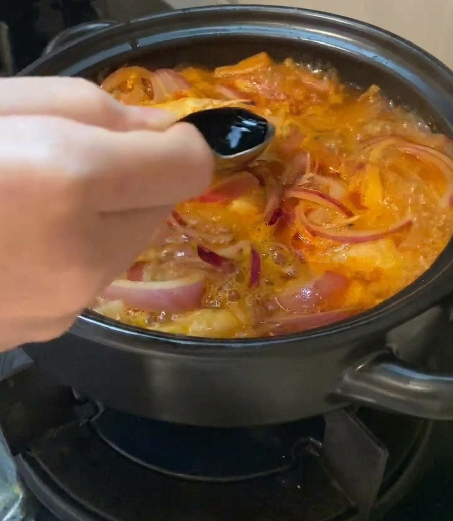 Ręka trzymająca metalową łyżkę i wlewająca ciemny sos sojowy do bulgoczącej zupy kimchi z cebulą.