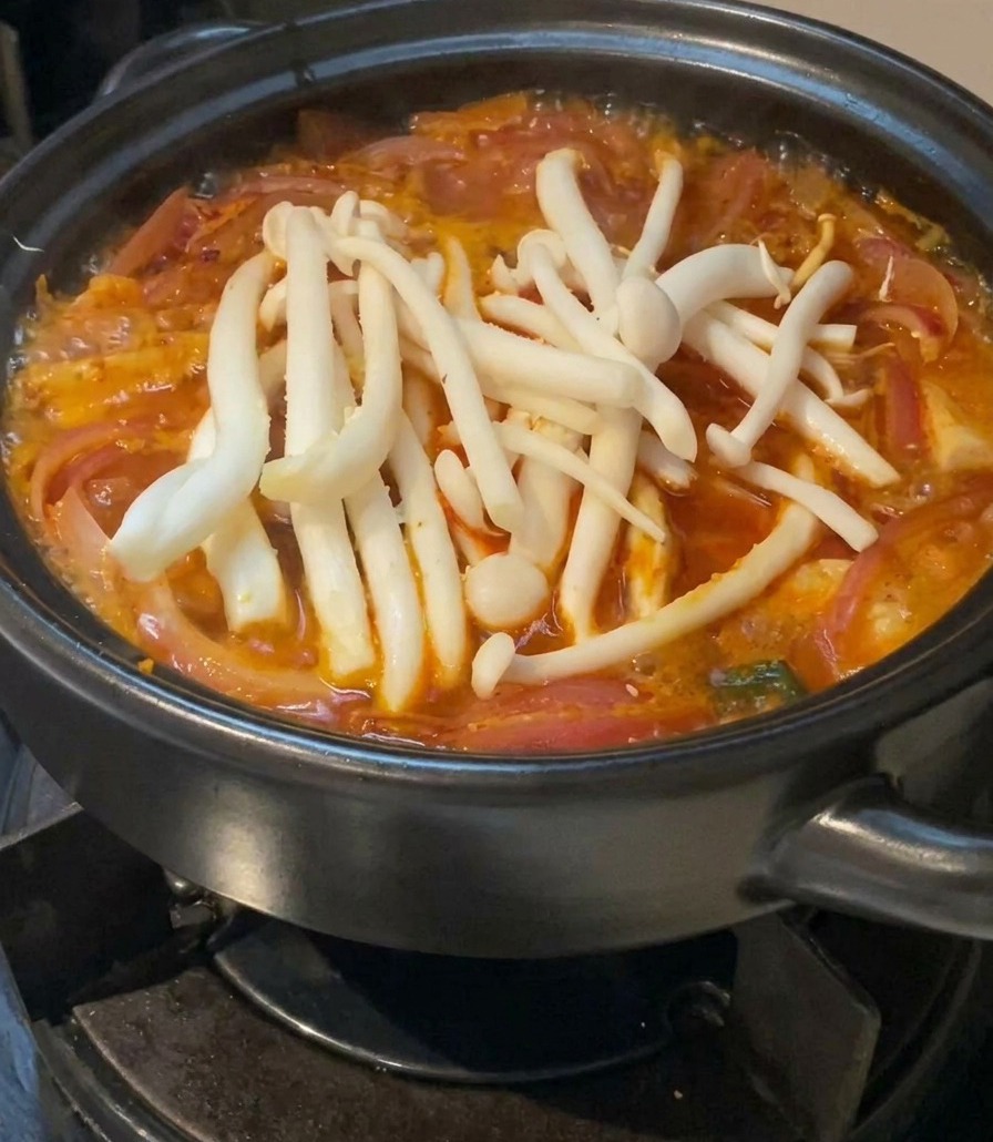 Kępa świeżych, białych grzybów shimeji ułożona na wierzchu czerwonej, gotującej się zupy kimchi w czarnym garnku.