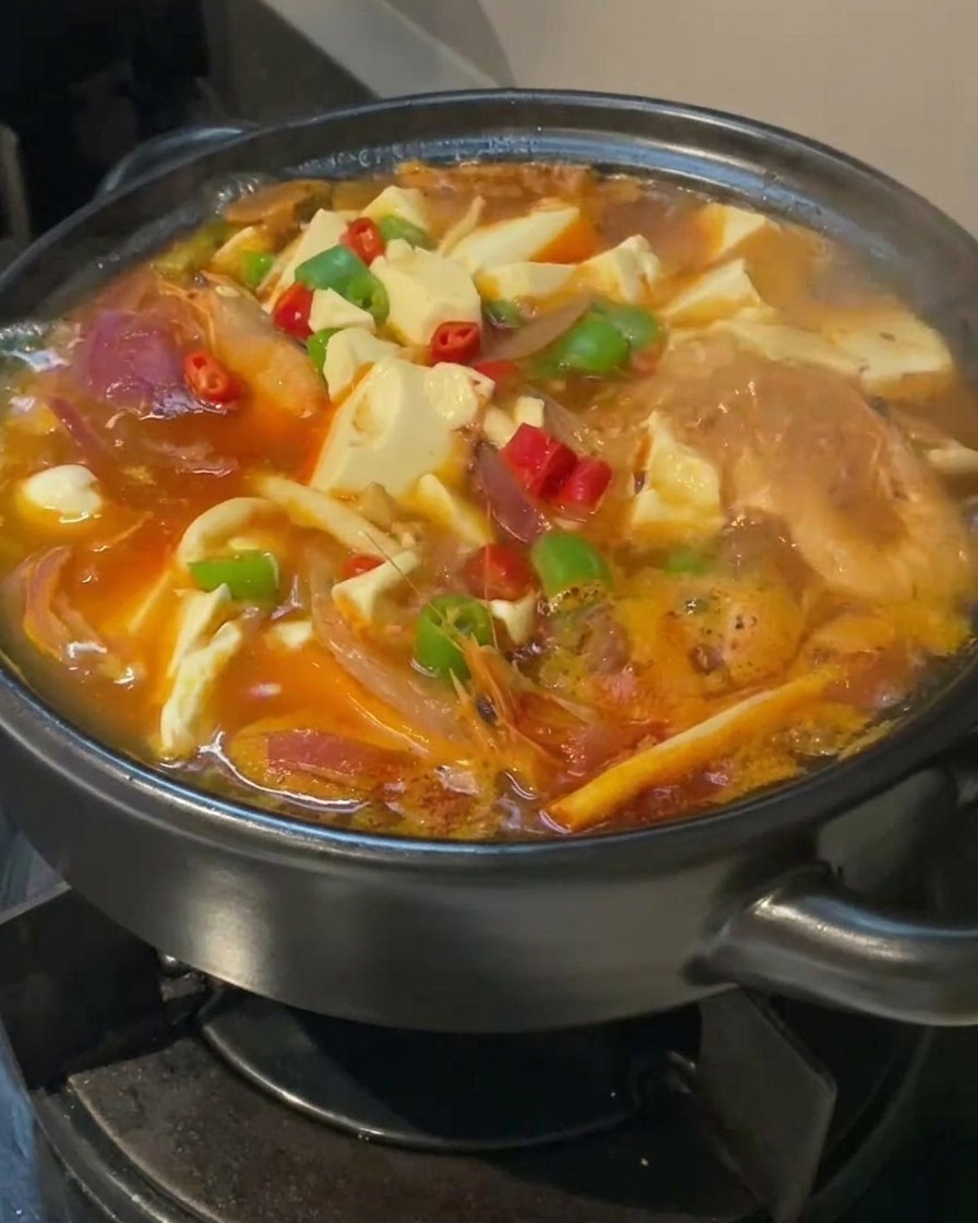 Zbliżenie na zupę kimchi z tofu wolno gotującą się w czarnym garnku, udekorowaną czerwonymi i zielonymi papryczkami.
