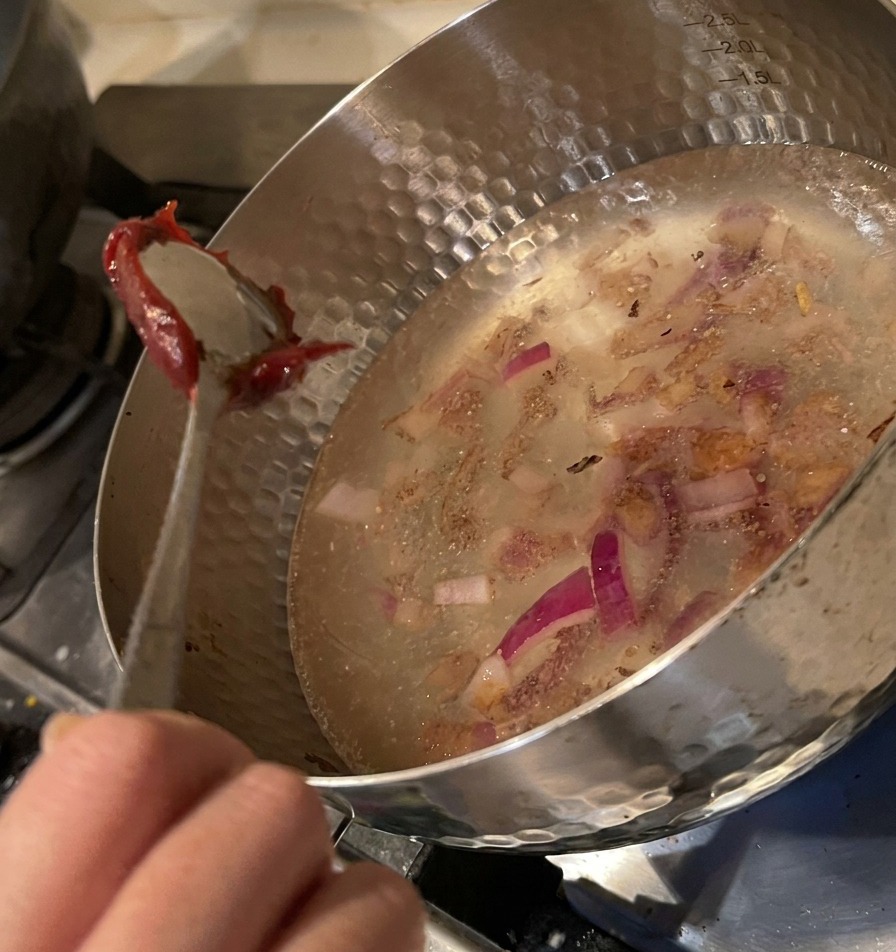 Łyżka nakładająca porcję gęstej czerwonej pasty chili do garnka z wodą i cebulą.