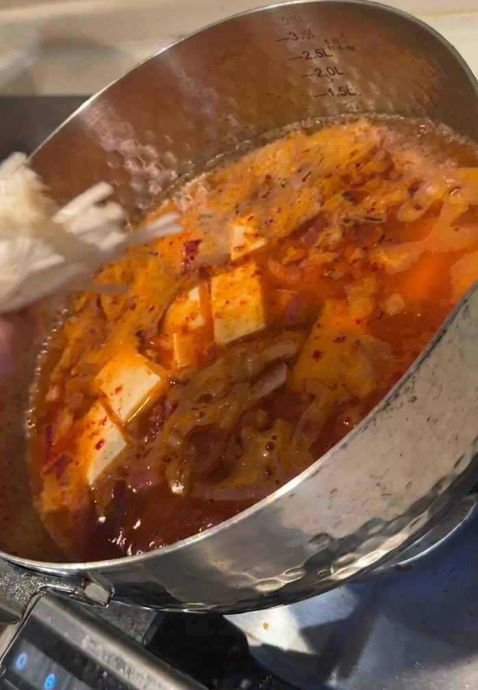 Ręka wkładająca kępkę białych grzybów enoki do garnka z bulionem i kostkami tofu.