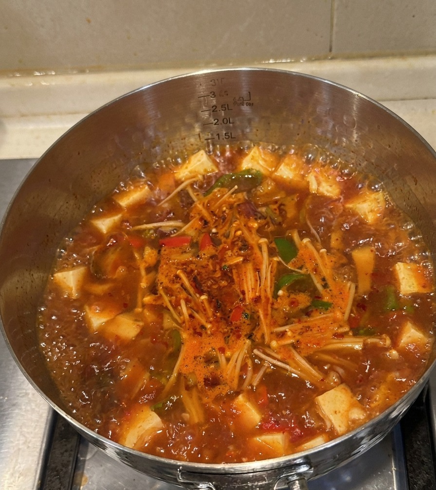 Garnki z gotującą się czerwoną zupą kimchi, widoczna ugotowana wołowina, grzyby i papryka.