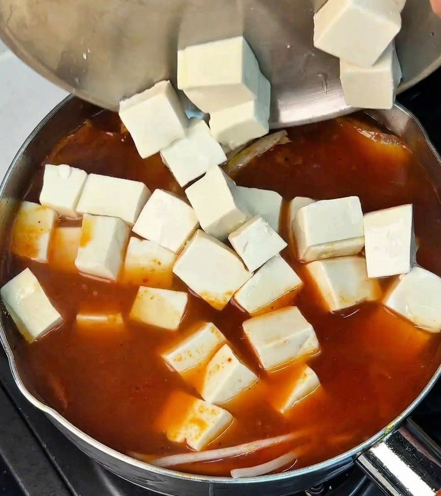 Kostki białego tofu wkładane do garnka z czerwonym bulionem.