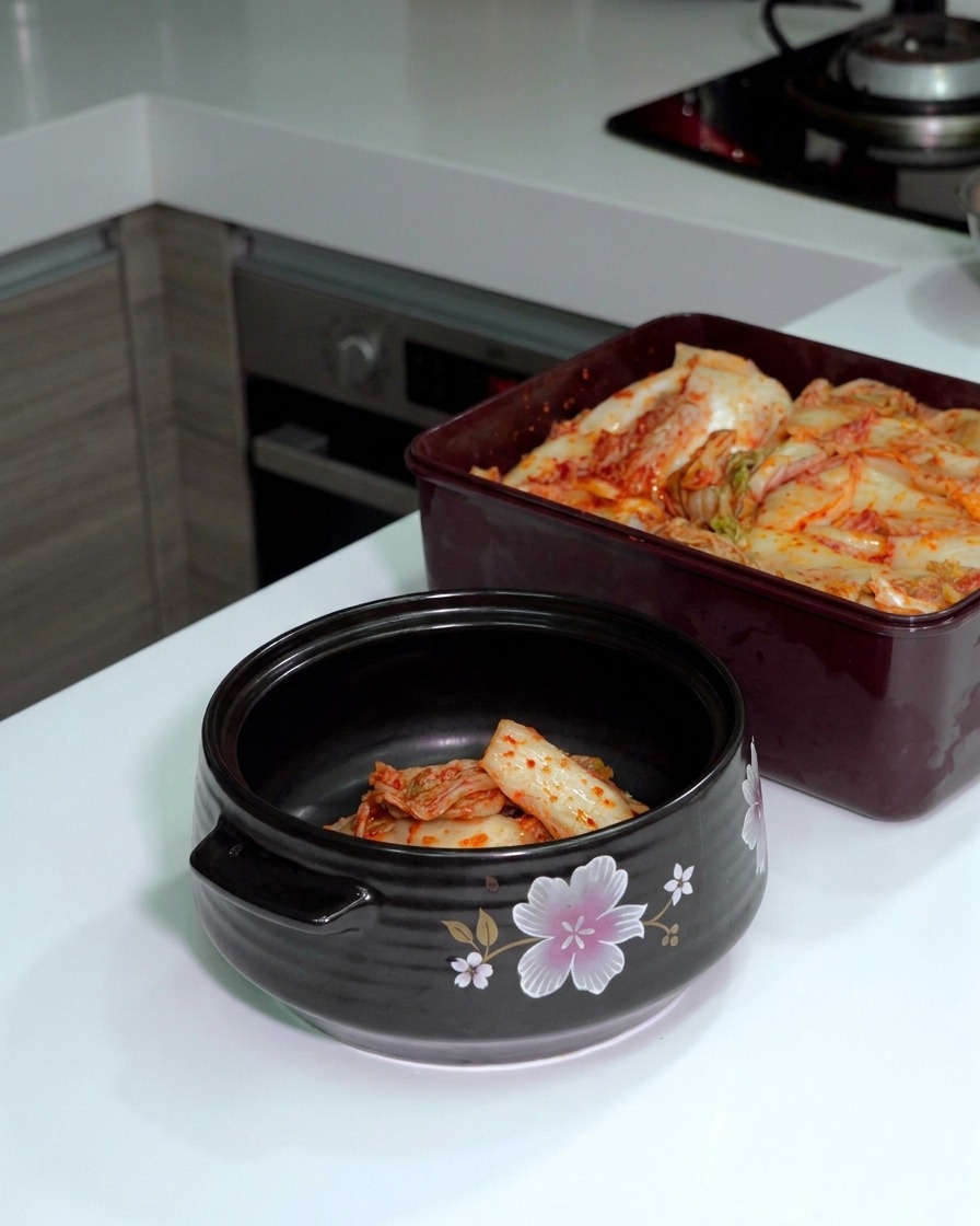 Sfermentowana kapusta kimchi krojona srebrnymi nożyczkami wewnątrz ciemnego ceramicznego garnka.