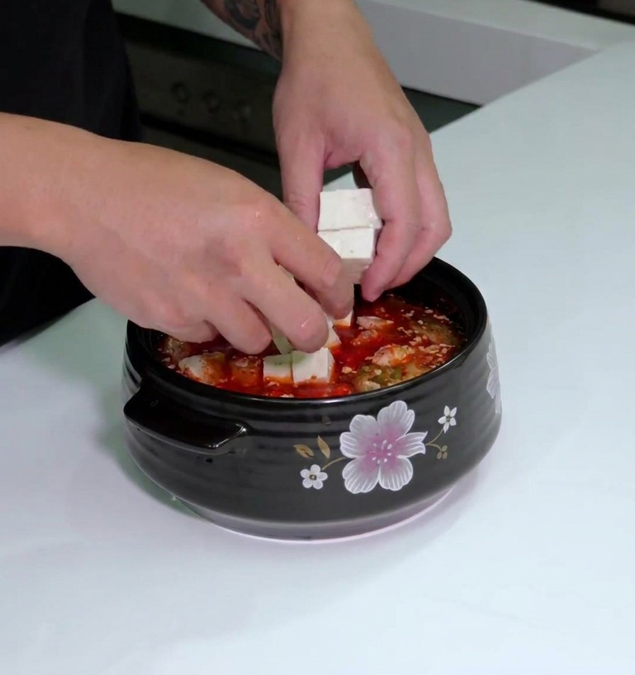 Dłonie kładące białe kostki tofu do garnka z czerwoną bazą kimchi i plastrami boczku.