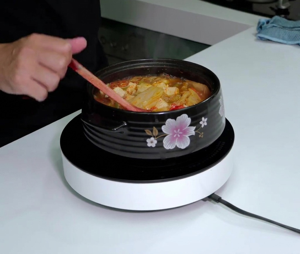 Butelka sosu Tabasco nad parującym, gotującym się garnkiem czerwonej zupy kimchi podczas mieszania drewnianą łyżką.