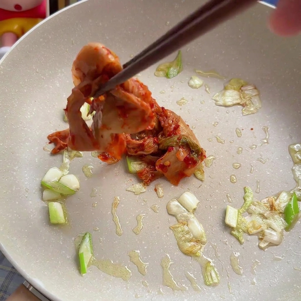 Pałeczki dodające porcję czerwonego, posiekanego kimchi do podsmażonej dymki na patelni.
