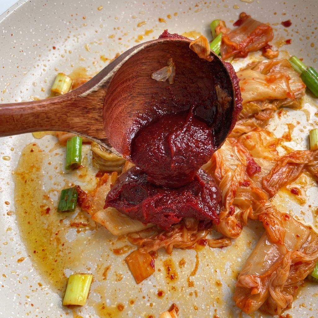 Drewniana łyżka nakładająca gęstą czerwoną pastę gochujang na smażone kimchi i dymkę na patelni.