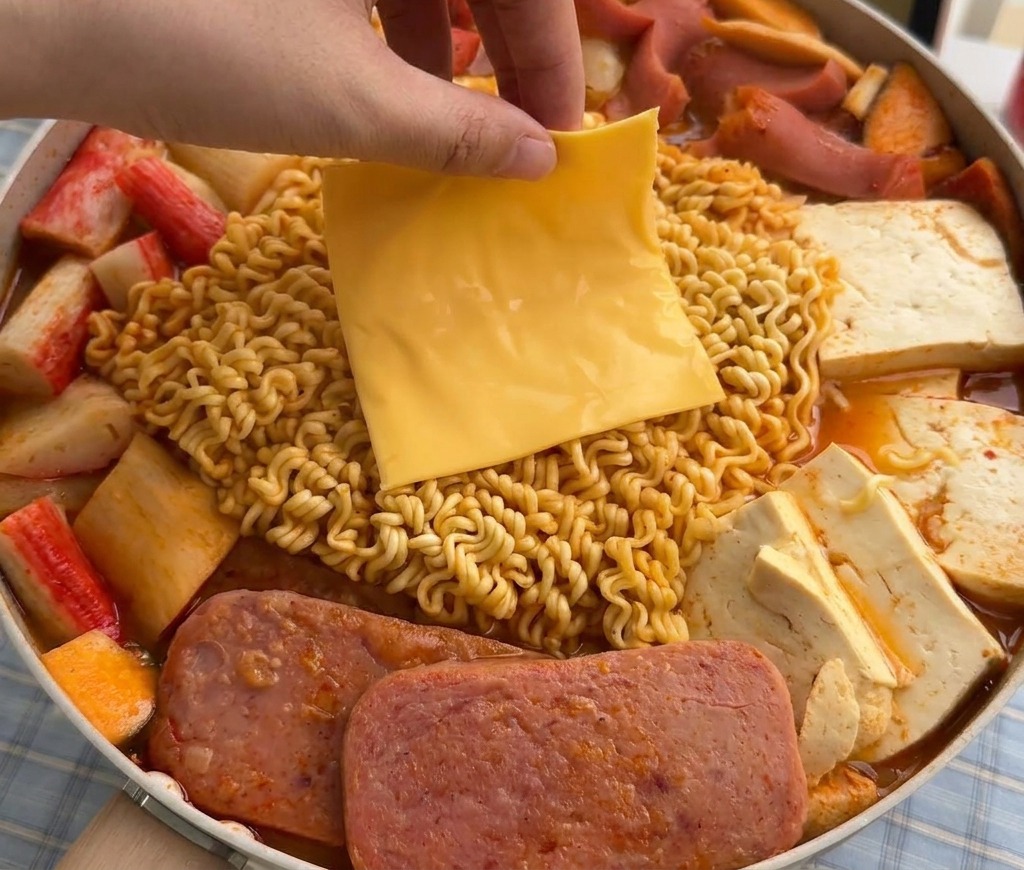 Dłoń kładąca plaster żółtego sera na mięknący makaron ramen w parującym garnku zupą wojskową.