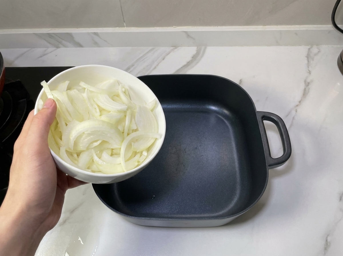Ręka trzymająca miseczkę z pokrojoną cebulą nad czarnym, kwadratowym garnkiem.