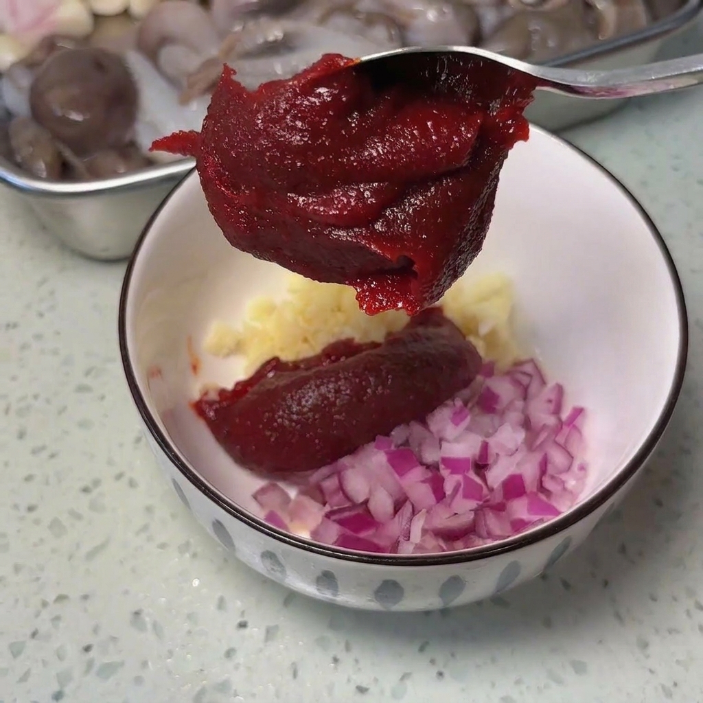 Metalowa łyżka z gęstą, ciemnoczerwoną pastą chili nad białą miską z czosnkiem i cebulą.