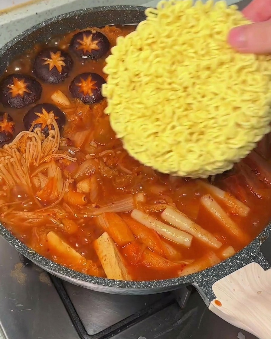 Dłoń wkładająca suchy makaron ramen do garnka z wrzącą, czerwoną zupą.