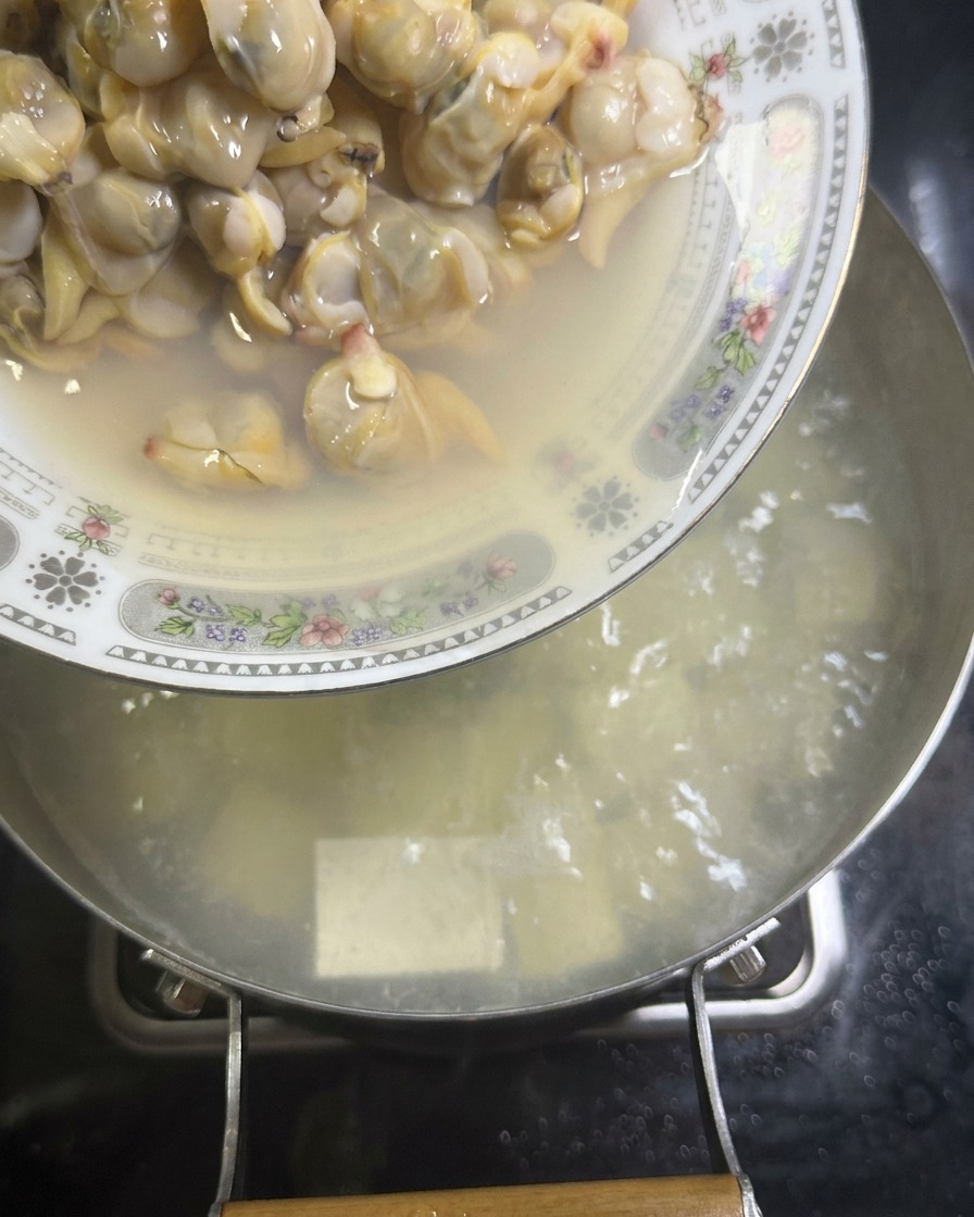 Talerz ugotowanego mięsa z małży jest wsypywany do garnka z gotującym się białym bulionem i kostkami tofu.