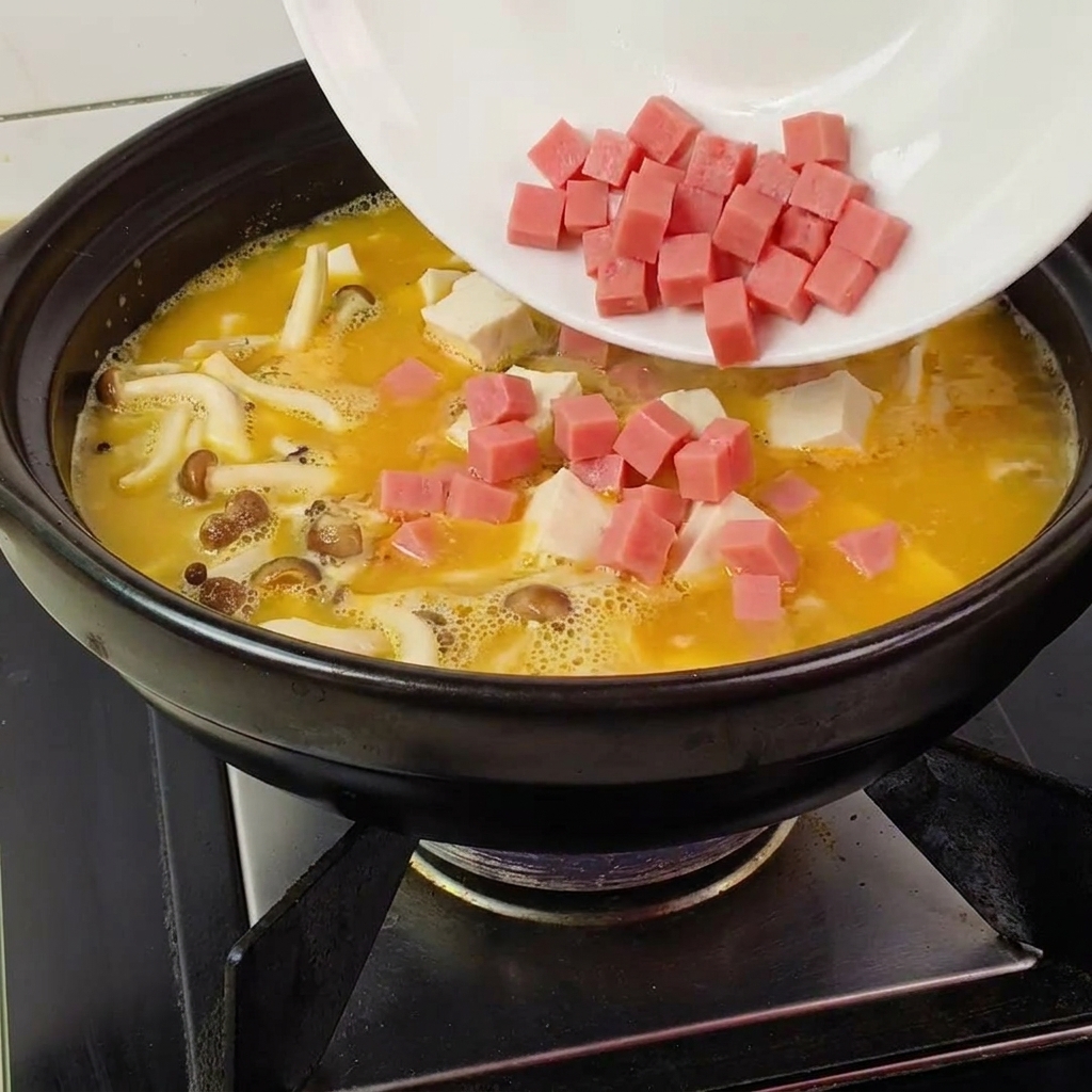 Kostki szynki wsypywane z talerza do garnka z żółtym bulionem, tofu i grzybami.