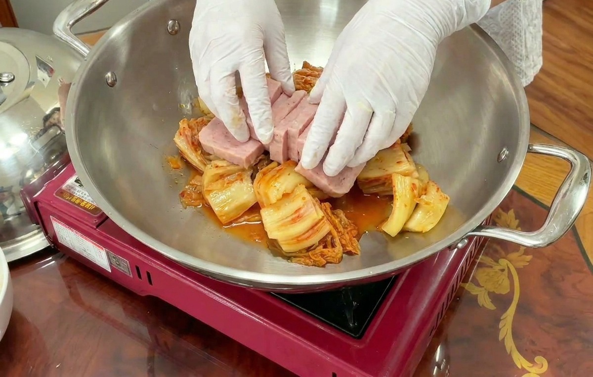 Dłonie w rękawiczkach starannie układają grube, prostokątne plastry różowej mielonki na warstwie kimchi w woku ze stali nierdzewnej.
